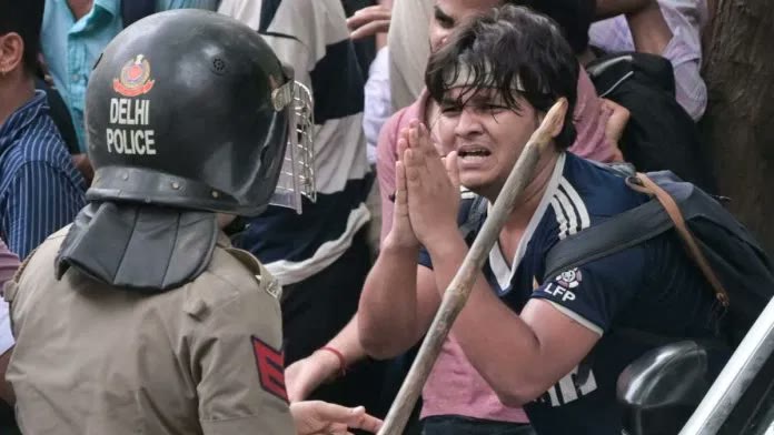 A young protester pleads with folded hands before a helmeted officer holding a lathi